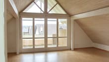 Spacious attic room with vaulted ceiling, wooden paneled walls, large windows providing ample natural light, and access to a balcony or terrace.