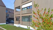 3-story modern building with wooden siding, large windows, and a balcony. The building is surrounded by a grassy area with decorative plants and shrubs.