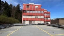 A multi-story red and white building with a large parking lot in the foreground. The building appears to be a commercial or industrial structure surrounded by trees and a cloudy blue sky.