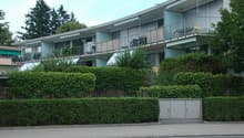 Two-story apartment building with balconies and awnings, surrounded by bushes and trees