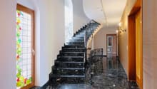 The image shows a grand staircase with black marble steps and an ornate wrought-iron railing. The walls are white, and there is a stained glass window with colorful geometric patterns. The hallway has a polished marble floor and wooden doors leading to ot