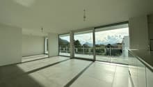 Spacious open-plan living area with large windows and sliding glass doors leading to a balcony or terrace, providing a scenic view of the surrounding mountains and landscape. The room features a minimalist design with light-colored tile flooring and white