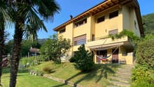 2-story yellow house with wooden roof, balconies, surrounded by lush greenery and palm trees, raised ground floor with steps leading to the entrance
