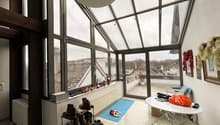 Room with large windows, a white table, a rug, and a shower head on the ceiling. The view outside the windows shows a snowy rooftop and cityscape.
