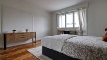 Bedroom with wooden floor, white walls, bed, wooden side table, desk, chair, window with curtains, rug, and potted plants.