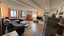 Wooden floor, beige walls, large windows, fireplace, living room couch, coffee table, TV, side table, rose in vase, desk, flower vase, basket on shelves, decorative items on shelves
