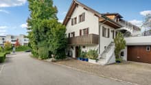 Detached house, 3 floors, white painted walls, brown roof, brick driveway, wooden garage door, brown balcony, plants, shrubs
