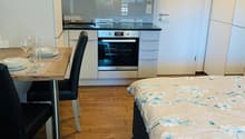 The image shows a studio apartment with an open-plan layout. The kitchen area features white cabinets, a built-in oven, and a dishwasher. There is a dining table with chairs near the kitchen. The living area has a bed with a floral patterned bedding. The