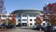 A modern, multi-story office building with a curved, domed roof structure. The building has large windows and is surrounded by trees with red autumn foliage. There is a parking lot in front of the building with several cars parked.