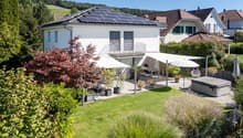 A modern white house with a roof-mounted solar panel system, a spacious backyard with outdoor seating and a jacuzzi, surrounded by greenery.