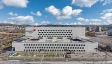 Large modern building with a distinctive white facade featuring a repeating pattern of circular openings. The building appears to be situated in an urban area with a view of the surrounding cityscape. There is a green landscaped area in the foreground of