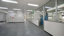 Empty office space with glass walls, white walls, gray floor, fluorescent ceiling lights, and a blue cabinet.