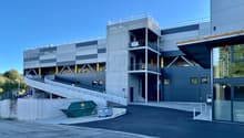 A large, modern commercial building with a gray and blue exterior. The building has multiple floors and a raised platform for loading and unloading. There is a dumpster visible in the foreground, indicating this is likely a storage or warehouse facility.