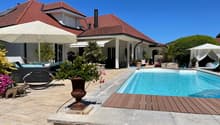 Swimming pool with wooden deck, surrounded by brick paving, patio with lounge chairs, and a large patio umbrella. There are also plants and flowers, and a wooden lounge chair with a canopy on the right side.