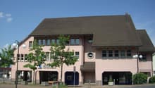 A two-story pink building with a slanted roof and large windows. The building has a sign that says 'Kirche Kantonal Bank' on the front. There are trees and a crosswalk in front of the building.