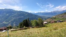 The image shows a scenic mountain landscape with rolling hills, lush green vegetation, and a small village nestled in the valley below. The sky is clear and blue, with wispy clouds. The mountains in the distance appear snow-capped. There is a fence or wir