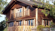 2 story wooden chalet, brown shingles, multiple glass windows, balcony with wooden railings, green trees in front