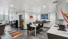 Open-concept living and dining area with modern, minimalist decor. The space features a large TV and entertainment system, a dining table with chairs, and a kitchen area with a dishwasher. The walls are decorated with colorful artwork, and the room has a