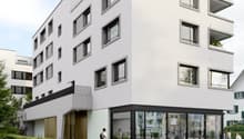 Modern white building, multiple windows, ground floor with glass walls, balcony on the second floor, greenery around