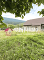 House with a brown roof, two windows, and a white wall, situated in a grassy area. A watermark from 'SAINT-IMMO' is displayed in the center of the image, with another house in the background and a mountain in the distance. The sky is clear and blue.
