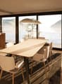 Spacious open-plan living and dining area with large windows offering a scenic view of the lake and mountains. The room features a wooden dining table with chairs, a kitchen area with built-in appliances, and a cozy seating area with a sofa. The overall d