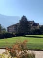 A wide grassy area with well-maintained trees and bushes, leading to a building complex with apartments in the background. Mountains can be seen in the distance.