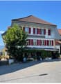 2 story house, white painted walls, red shutters, brown roof, outdoor seating area with trees and plants, covered entrance with awning, several steps leading up to the entrance