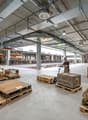 large warehouse with many wooden pallets, boxes, and items, a person using a pallet jack, high ceilings, metal beams, and lighting