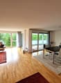 Spacious open-plan living and dining area with large windows overlooking a balcony or terrace. The room features hardwood floors, a brown leather sofa, a coffee table, and a dining table with chairs. There is also a media console and shelving unit against
