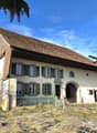 atypical house with tiled roof and raised ground floor, white walls with green shutters, green fence, driveway in front