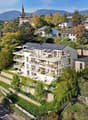Modern white house with solar panels, multiple balconies, surrounded by trees and greenery, with a view of the mountains