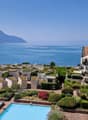 This image shows a scenic apartment complex overlooking a beautiful lake surrounded by mountains. The complex features a series of terraced buildings with balconies and patios, as well as a large swimming pool in the foreground. The buildings have a Medit