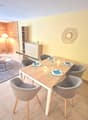 The image shows a combined living room and dining area. The living room has a wooden accent wall, a round decorative wall hanging, and a coffee table. The dining area has a wooden table with four gray upholstered chairs, and a sideboard unit. The room has