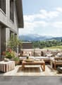 Spacious outdoor patio with wooden deck, comfortable seating area with sofa and armchairs, coffee table, decorative plants and flowers, and scenic mountain view in the background