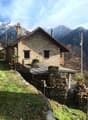 a stone house with a wooden front porch, elevated ground floor, chimney, windows, flag, and lush greenery, nestled against a mountainous backdrop