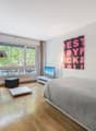 A room with a bed, a couch, a TV, a lamp, a table, and a window with a view of the outside.