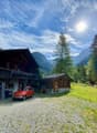 2 story wooden house, gravel driveway, steps leading to porch, red car, small wooden cabin, sunny weather, mountains in the background