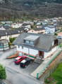 Aerial view of a small town or village nestled in a mountainous, forested landscape. The image shows a cluster of residential buildings with tiled roofs, some with balconies or terraces. There are several parked cars visible in the foreground, indicating