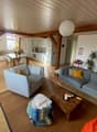 Open living room with wooden floor, wooden beams, white walls, two sofas, wooden coffee table, two armchairs, carpet, dining table, chairs, kitchen, microwave, refrigerator, potted plants, shelves, decorative items