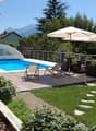 Outdoor swimming pool surrounded by patio furniture and umbrellas, with a grassy lawn and landscaping visible.