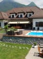Large two-story house with a tiled roof, surrounded by mountains. The house has a patio with a swimming pool, wooden furniture, and a stone wall. There are also parking spaces on the property.