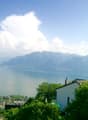 The image shows a scenic view of a lake surrounded by mountains and lush greenery. The lake appears to be calm and reflective, with buildings and houses visible along the shoreline. The sky is clear with some fluffy white clouds, creating a picturesque an