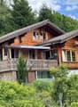 Two-story wooden chalet-style house with a balcony, surrounded by lush greenery and trees
