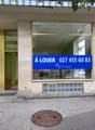 Exterior of a commercial building, white and gold concrete walls, glass entrance with brass frame, blue sign indicating 'A LOUER' with contact number.
