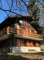 Two-story wooden chalet-style house with a balcony, surrounded by trees and a grassy area