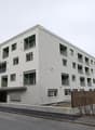 Multi-story apartment building with gray exterior, balconies, and windows. The building is situated in a mountainous area with other buildings and vehicles visible in the surroundings.