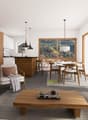 Open concept living and dining area, gray tile floor, glass windows, wooden coffee table, couch, chair, TV, dining table and chairs, kitchen island, large wooden cabinet, kitchen countertop with stools, potted plants