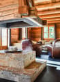 Rustic log cabin interior with wooden walls and ceiling, stone fireplace, leather armchairs, and a coffee table on a tiled floor