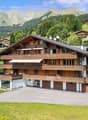 Large wooden chalet, multiple balconies, garage, set in a mountainous area with green slopes and pine trees.