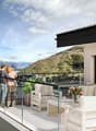 large balcony with plants, two people standing on balcony, white couches, table with flower vase, glass railing, mountain view in the distance